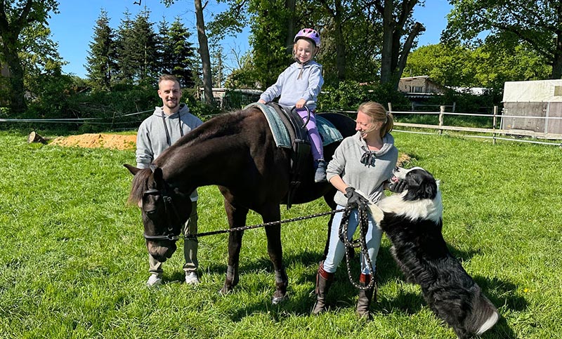 Ein Mädchen sitzt auf einem Pferd. Ein Mann und eine Frau mit Hund stehen daneben.