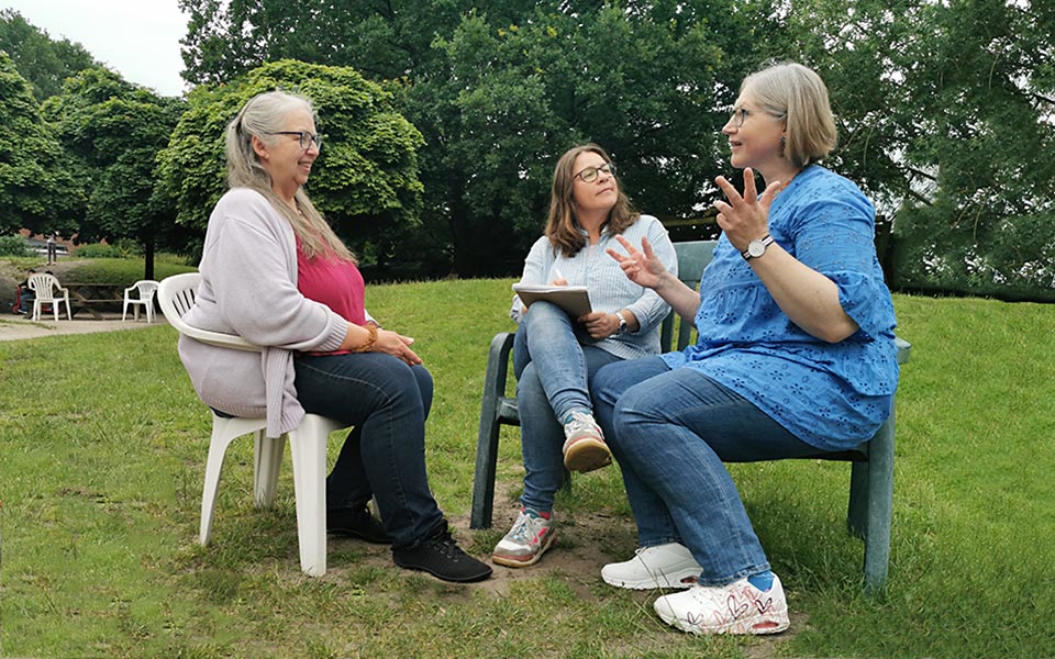 Drei Mitarbeiterinnen des Kompetenzteams sitzen draußen auf Stühlen und sprechen miteinander.