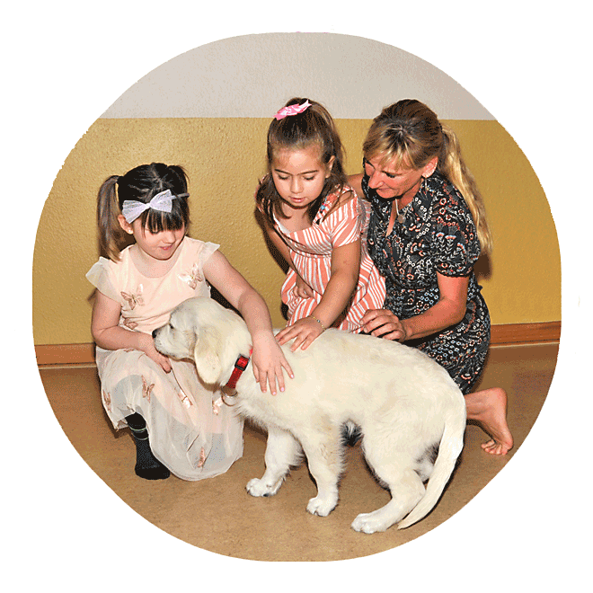 Zwei Mädchen streicheln einen Therapiehund, der an der Hand des einen Mädchens schnuppert. Eine Therapeutin macht den Mädchen Mut dabei.