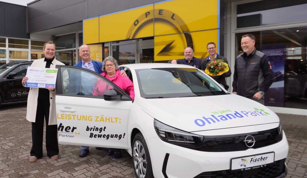 Sylvia Fabisch von der Lebenshilfe Kaltenkirchen hält den Scheck über 17.000 Euro in die Kamera. Neben ihr sind das Auto, die glücklichen neuen Besitzer und weitere Beteiligte zu sehen.